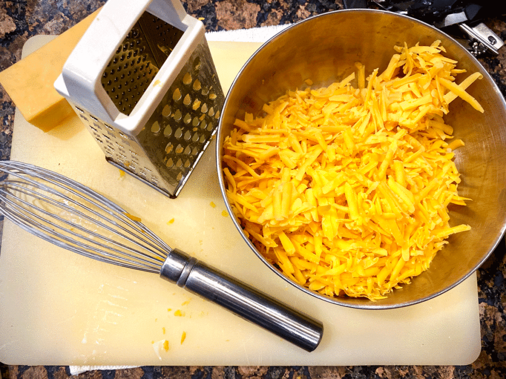 Grated Cheddar cheese in a bowl with a grater and whisk on a cutting board