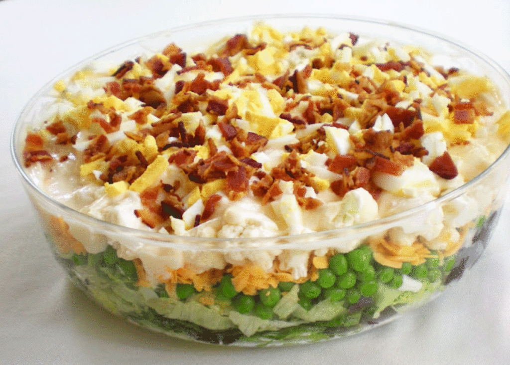 Bowl of layered salad with peas, cauliflower, cheese, dressing and bacon.