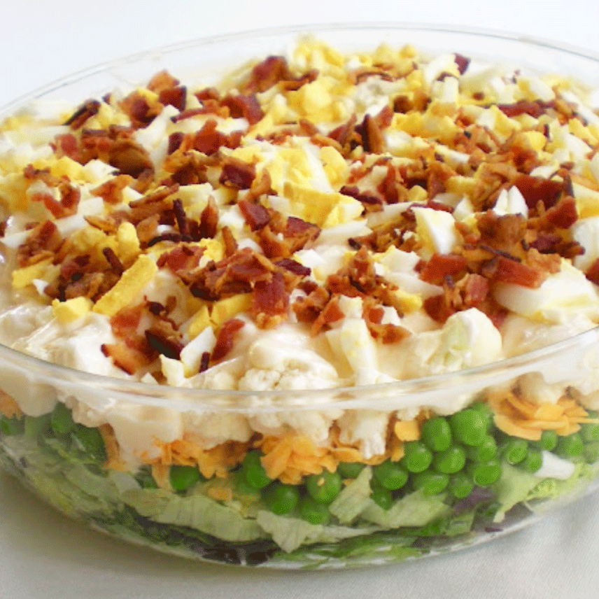 Bowl of layered salad with peas, cauliflower, cheese, dressing and bacon.