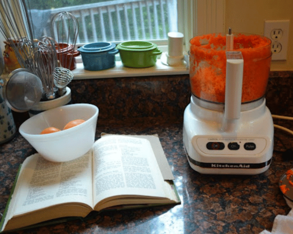 A food processor with shredded carrots on a kitchen counter with a cookbook propped open by a bowl filled with oranges