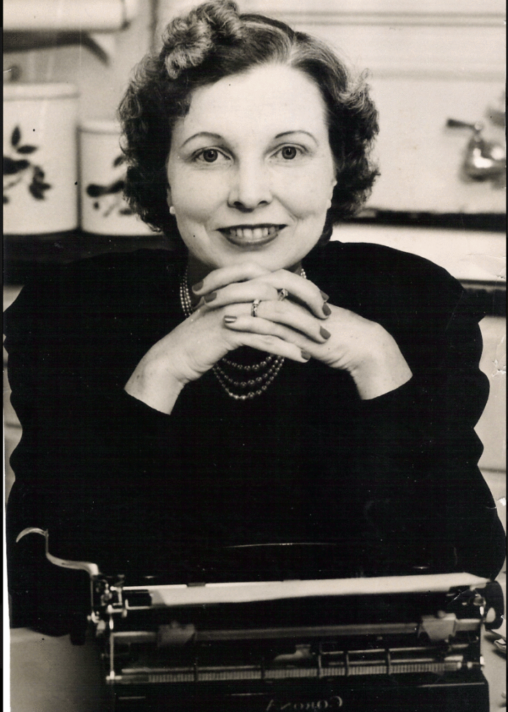 A black and white image of a woman sitting at a table with a typewriter with her chin on her folded hands